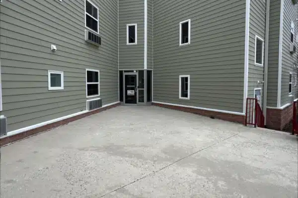 Before: Entrance to a building with a concrete area, surrounded by green siding and several windows. Red stairs on the right.
