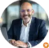 Smiling individual in a suit sitting at a laptop, with a blurred background of a modern office setting.