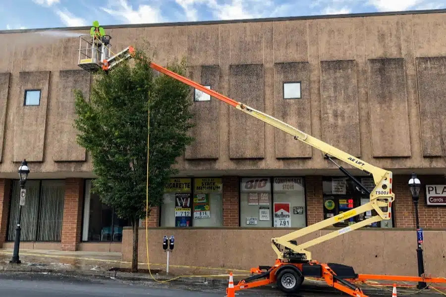A worker in a lift is cleaning the top of a building, with a tree beside the lift and shop signs visible below.