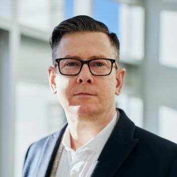 A person wearing glasses and a dark blazer stands in an indoor setting with large windows in the background.