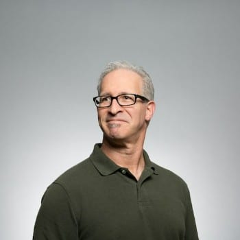 A smiling man in a green polo shirt, wearing glasses, stands facing slightly to the left against a light gray background.