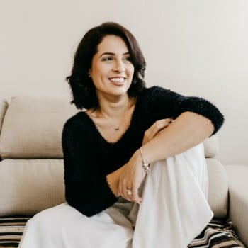 A person in a black sweater smiles while sitting on a couch with legs crossed, wearing white pants.