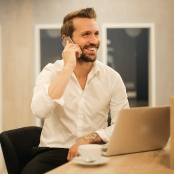 Smiling man in a white shirt talking on a smartphone, sitting at a desk with a laptop and a cup.