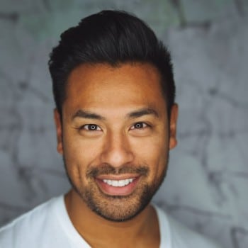 Smiling man with dark hair, wearing a white shirt, against a blurred background.